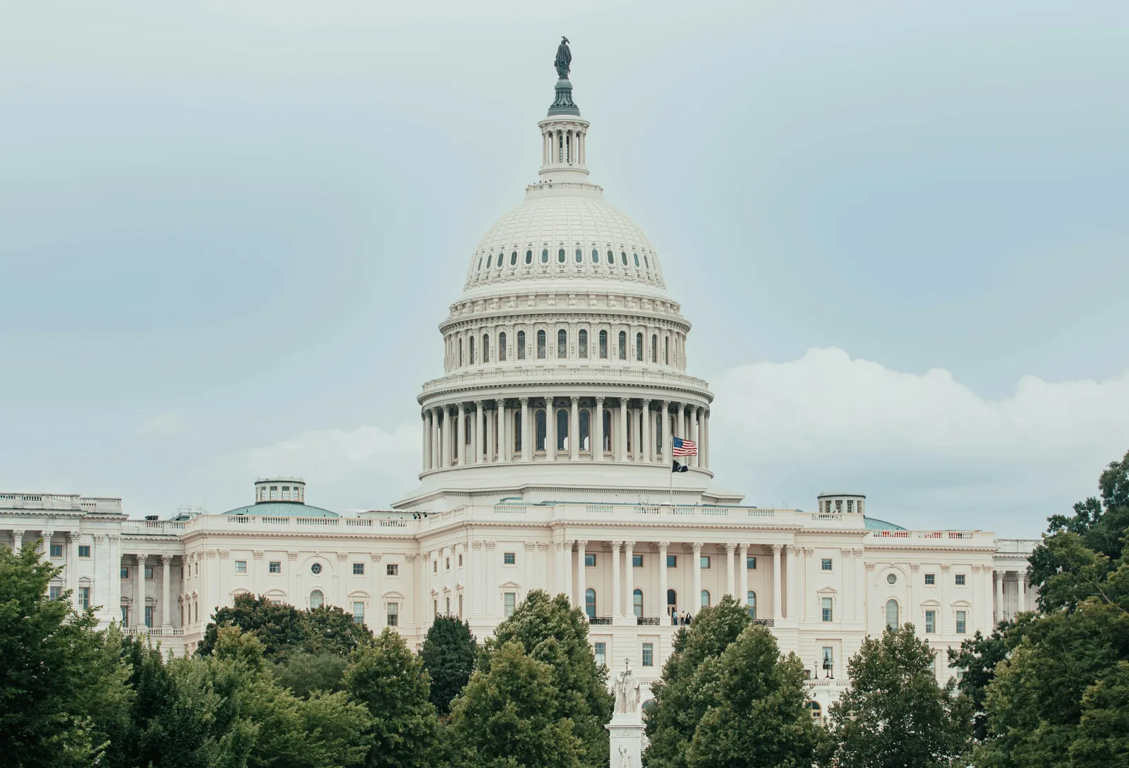 The United States Capitol building with its large dome, American flag, and surrounding trees under a cloudy sky.