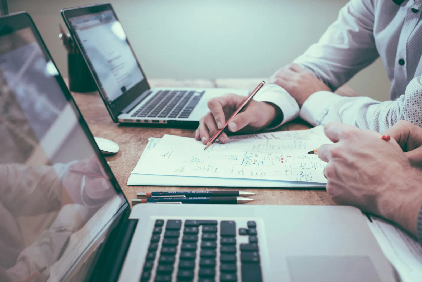 Two professionals collaborating over laptops and documents with handwritten notes at a wooden desk.