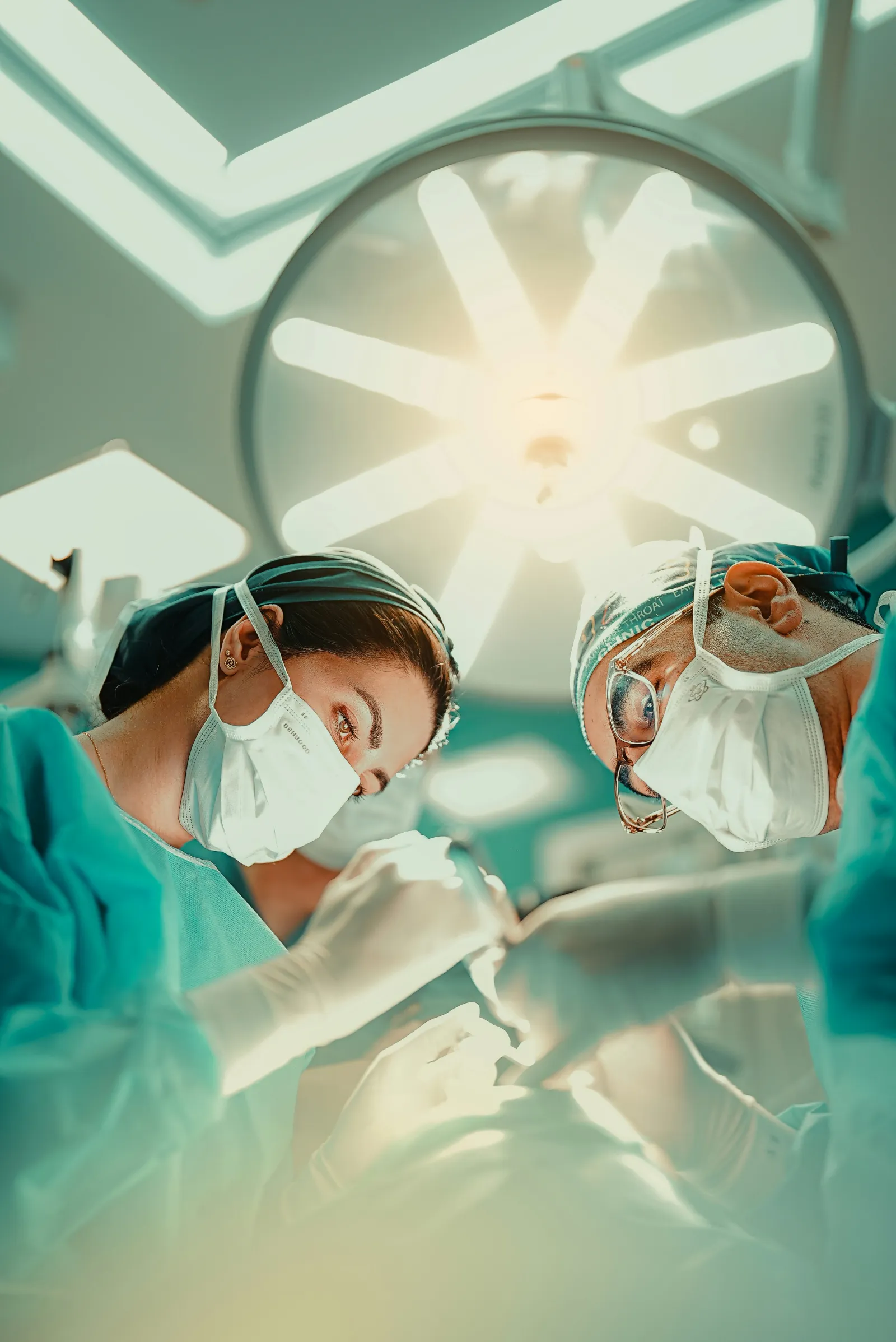 Two surgeons wearing masks and scrubs performing surgery under bright operating room lights.