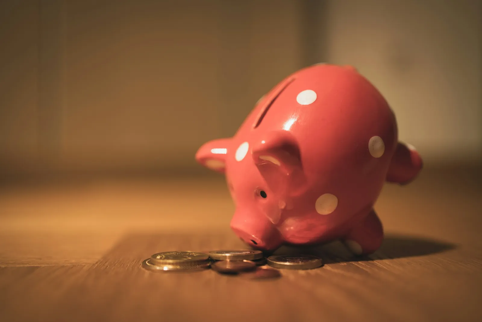 Close-up of a pink piggy bank with white polka dots tipped over beside scattered coins on wooden surface.